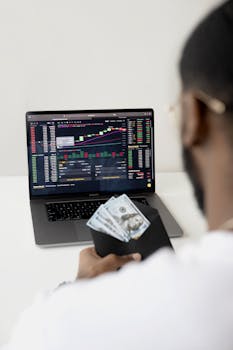 A person examines stock market data on a laptop while holding dollar bills, showcasing finance and trading concepts.