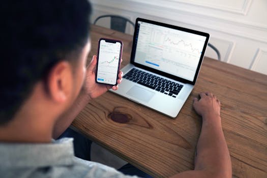 Individual reviewing stock market trends using a laptop and smartphone at a wooden table.